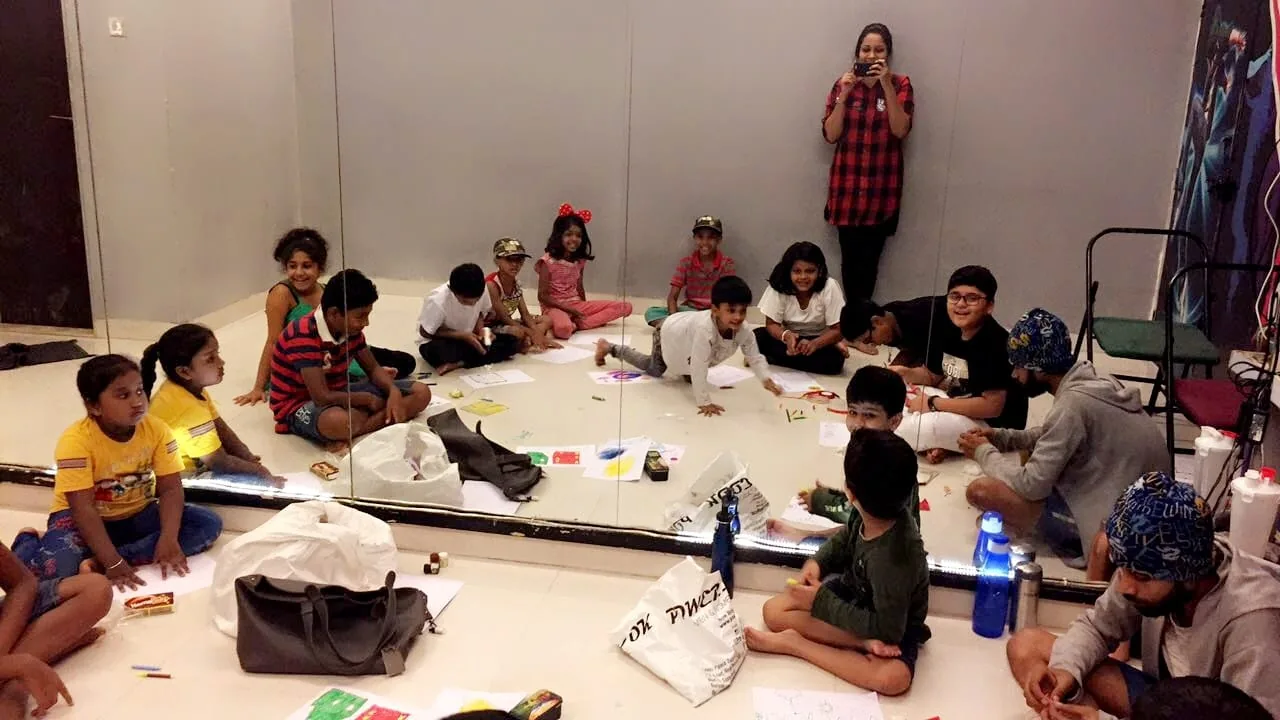 Children sitting in a circle on a studio floor drawing on paper with crayons