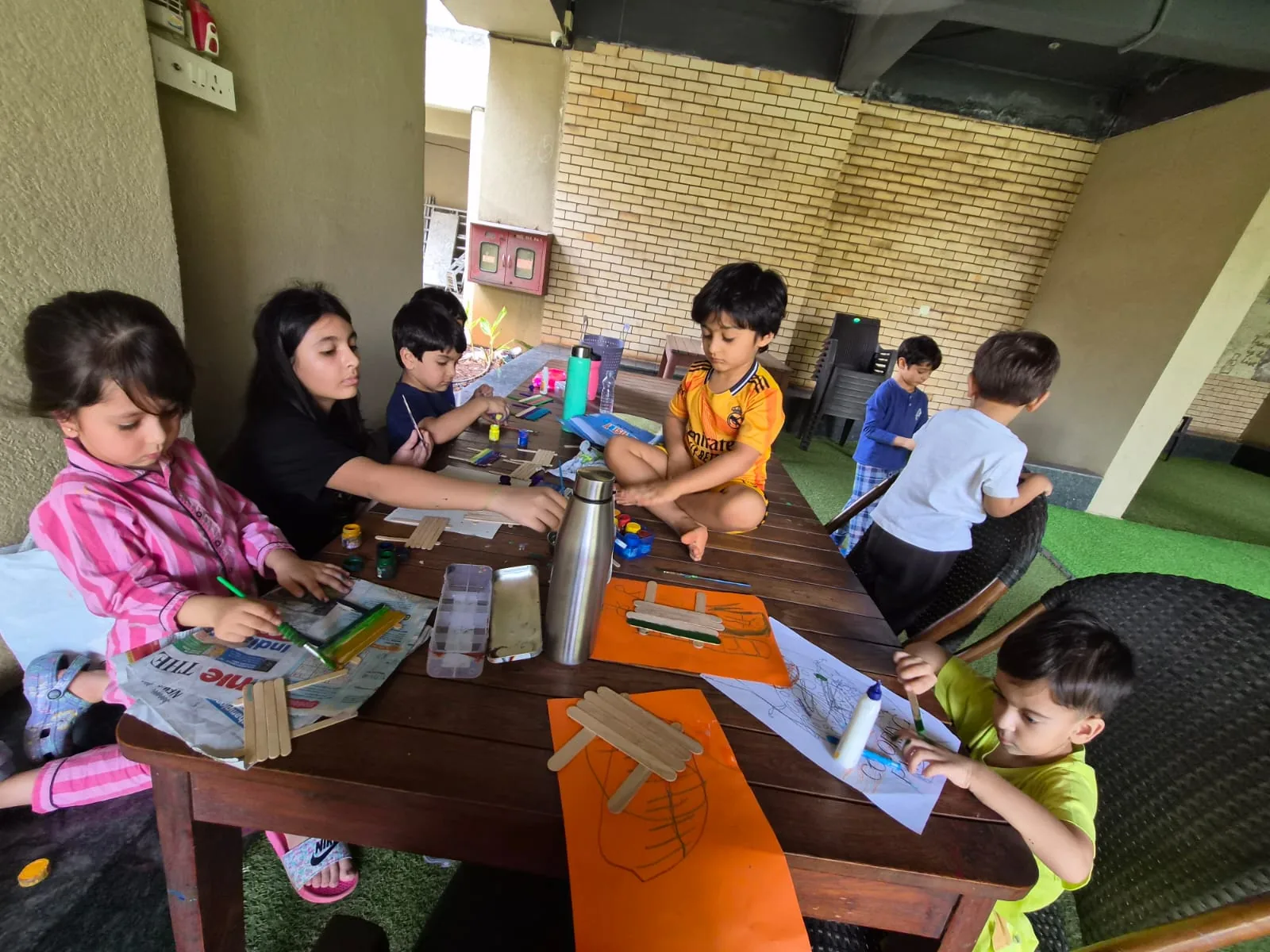 Children of different ages painting popsicle sticks together at a long wooden table on a balcony